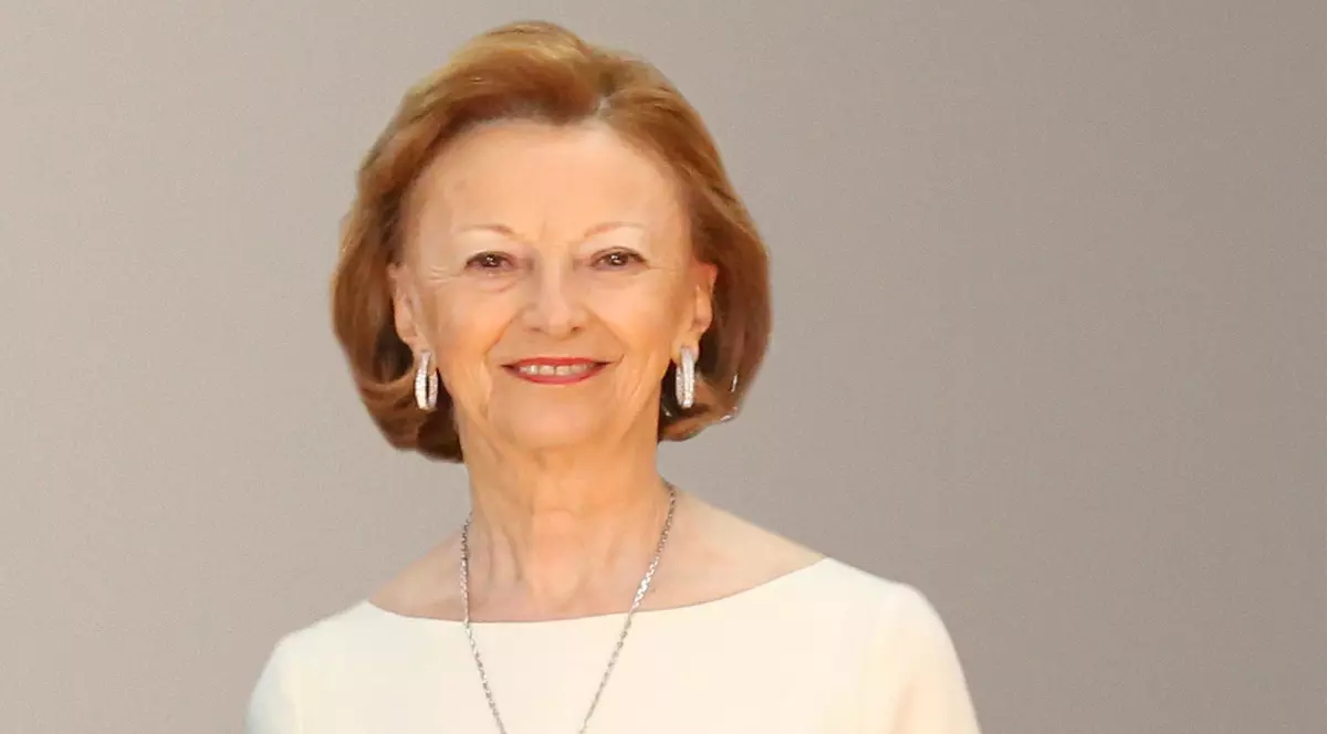 În imagine apare o femeie elegantă, îmbrăcată într-o rochie deschisă la culoare, cu bijuterii discrete și o expresie calmă și zâmbitoare. Portretul sugerează rafinament și sobrietate, fiind realizat într-un cadru simplu, cu fundal neutru.