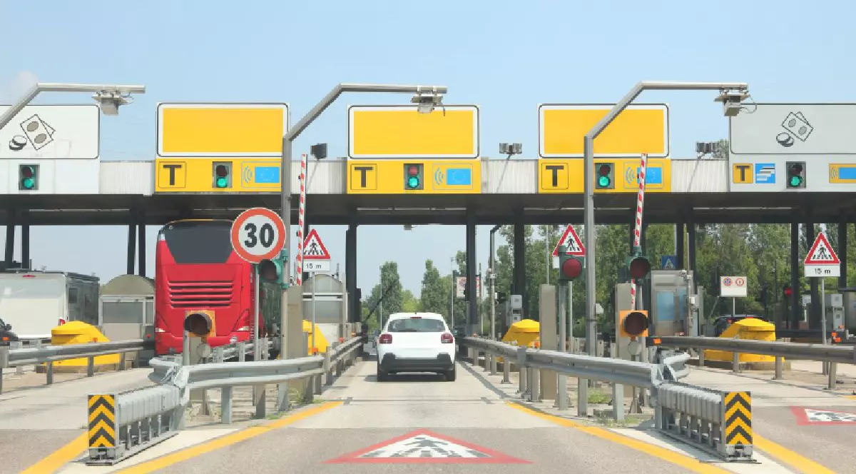 Fotografie cu mai multe cabine de taxare, indicatoare și semafoare, un autobuz roșu și o mașină albă oprite la bariere.