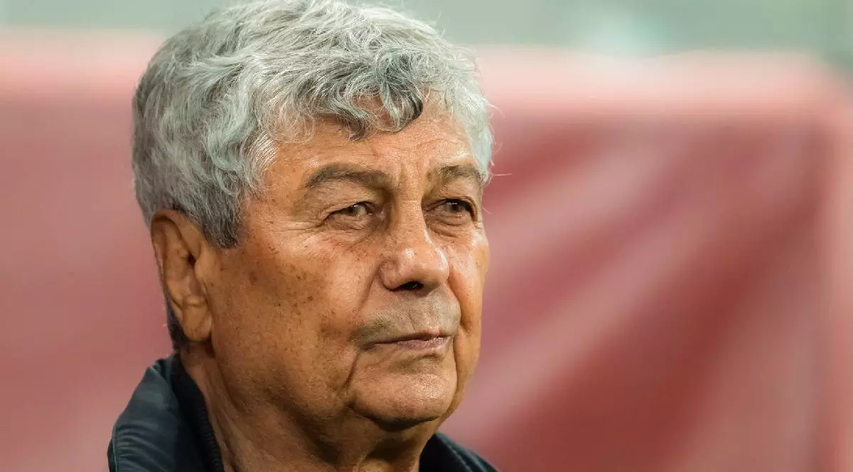Fotografie cu Mircea Lucescu, cu părul scurt, creț și cărunt, îmbrăcat într-o jachetă neagră, pe un fundal roșu estompat.