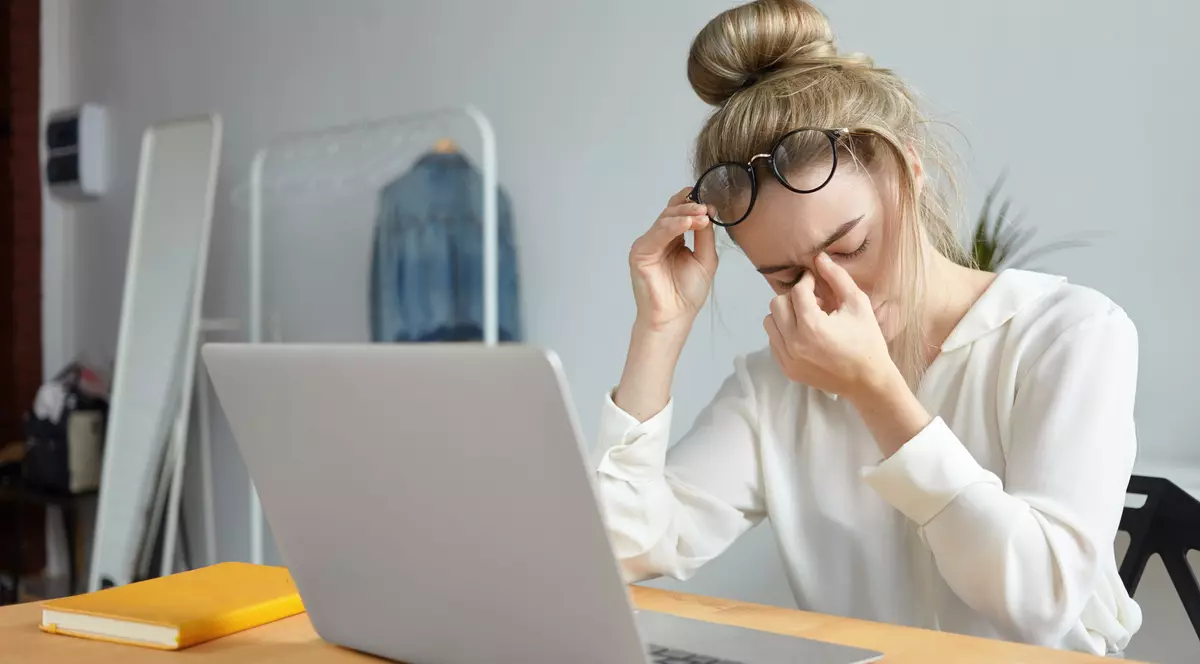 O femeie își ține mâna în jurul ochilor și cu cealaltă ochelarii, după o zi de muncă grea în fața calculatorului