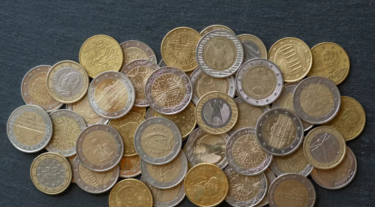 Imaginea prezintă un teanc de monede euro, de diferite valori, împrăștiate pe o suprafață închisă la culoare. Se pot observa atât monede de 1 și 2 euro, cât și piese mai mici, cu modele și simboluri specifice mai multor țări din zona euro.