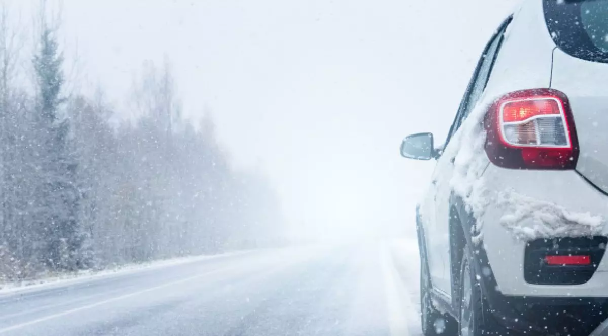 Această fotografie surprinde partea din spate a unei m așini albe, care circulă pe un drum acoperit de zăpadă, înconjurat de o pădure densă de copaci. Vehiculul este parțial ascuns de ninsoarea abundentă Drumul este acoperit de zăpadă, pe care se văd urmele roților.