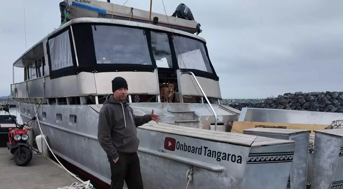Un barbat, cu caciula pe cap, hanorac gri si pantalon negru, in marina, arata spre iahtul Tangarao, barca pe care a renovat-o cu sotia sa