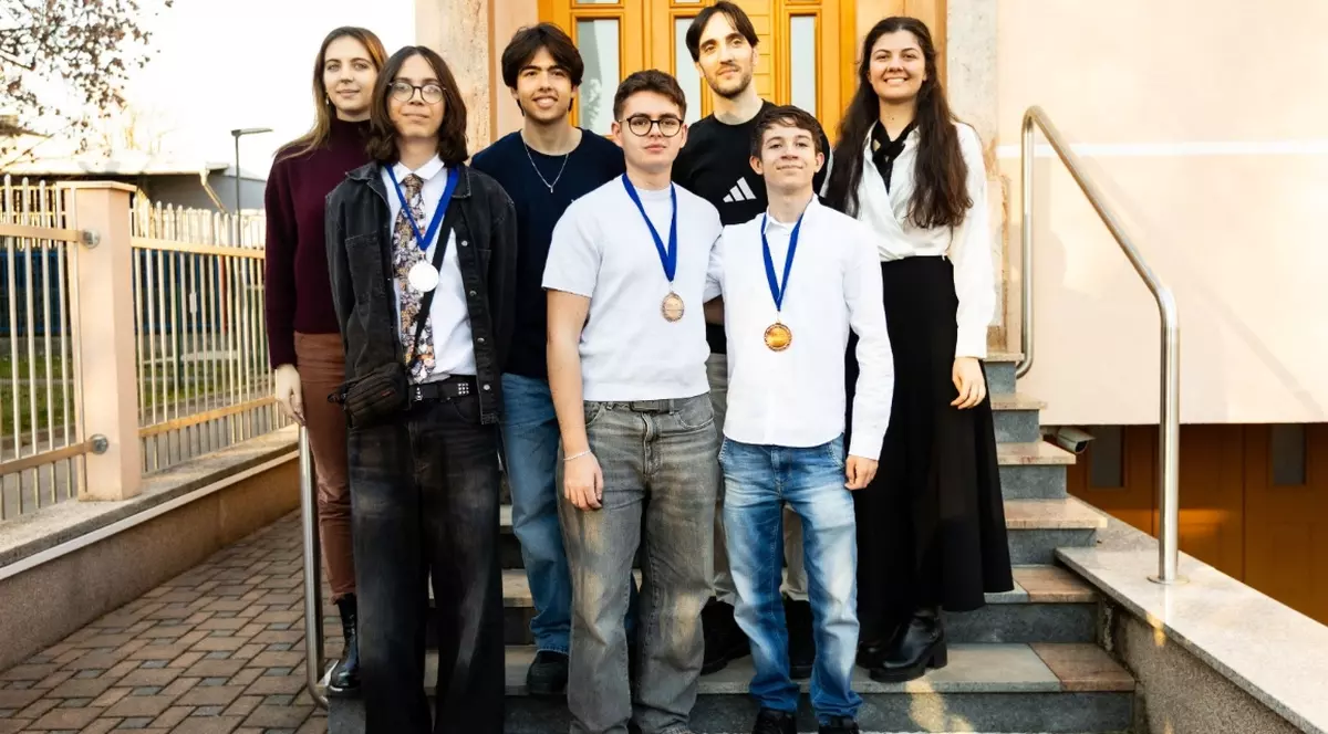 În imagine apar șapte tineri care pozează pe treptele unei clădiri, doi dintre ei purtând medalii la gât. Grupul este îmbrăcat casual-elegant și zâmbește, sugerând un moment de mândrie și celebrare a unei performanțe.