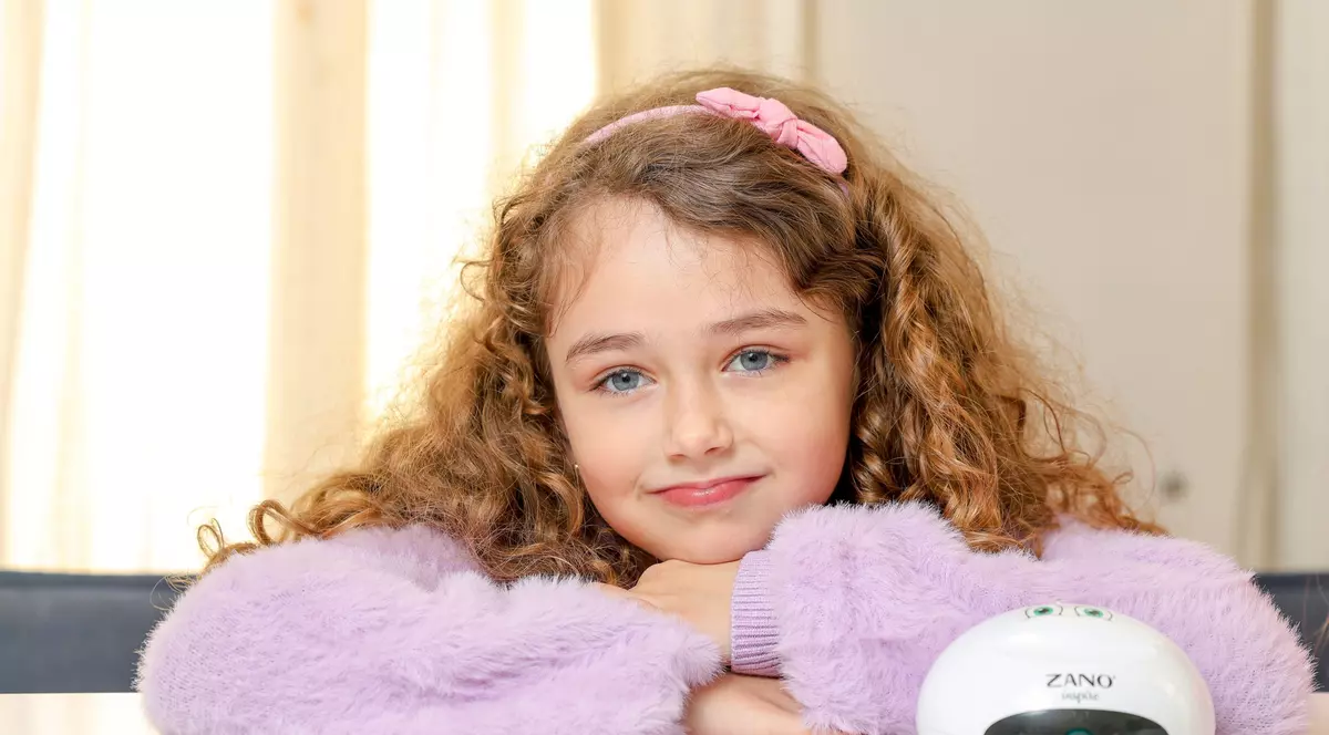 Fetita rosacata, cu par lung, cret si ochi albastri, sta rezemata cu barbia pe brate pe masa. In fata ei este un inhalator de la ZANO