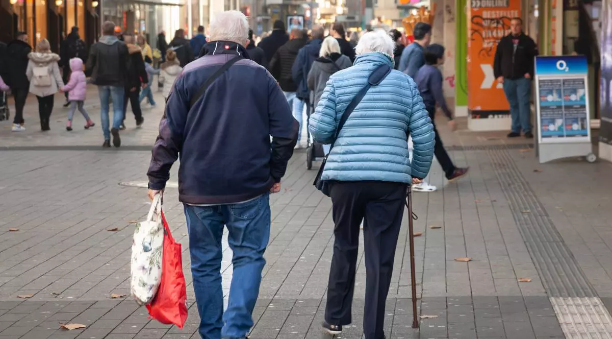 Imagine neclară selectivă a unui cuplu de bătrâni care merg pe stradă, bărbatul care două sacoșe, femeia folosește o cârjă, multi oameni în jurul lor
