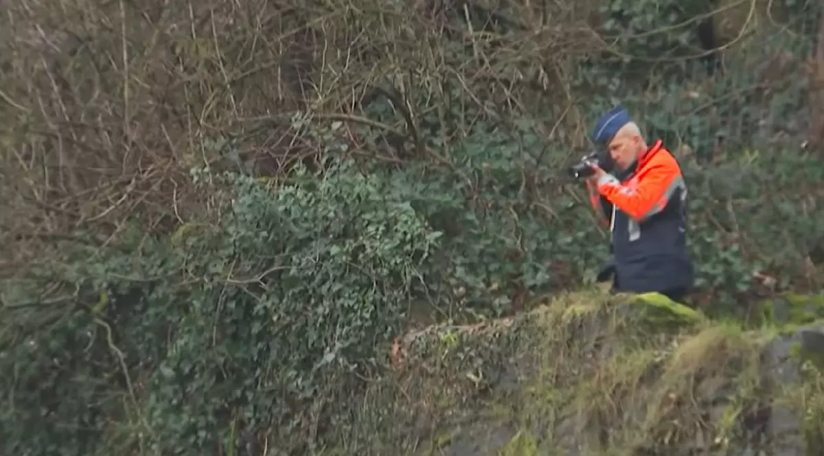 Un bărbat cu părul alb și chipiu albastru, îmbrăcat într-o jachetă portocaliu cu negru, înconjurat de vegetație și ținând în mâini un aparat foto.