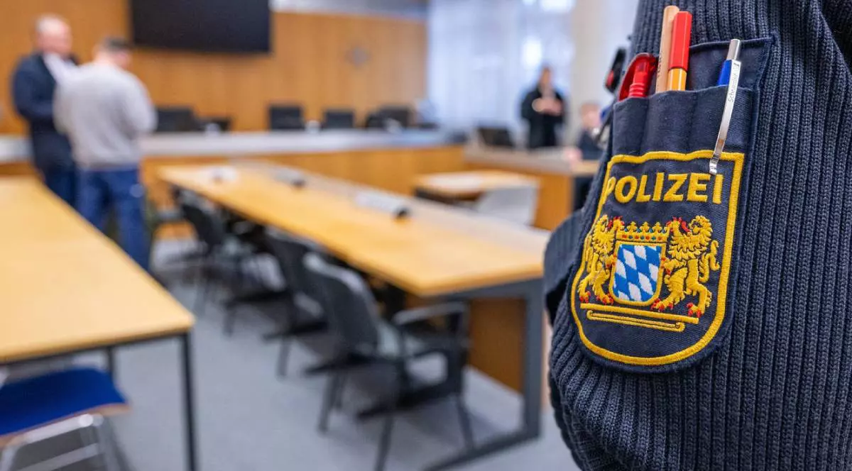 Un polițist cu insigna poliției bavareze stă în sala de judecată a tribunalului districtual Landshut. Se vad mese si scaune, in findal cateva persoane, backgroundul este estompat. Procesul unui bărbat acuzat de tentativă de omor.