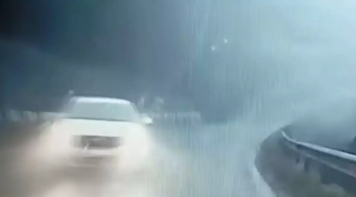 Imaginea surprinde o scenă pe un drum umed și încețoșat. O mașină albă se deplasează cu farurile aprinse dinspre stânga spre centru. Drumul face o curbă spre dreapta, cu un parapet vizibil pe partea dreaptă.