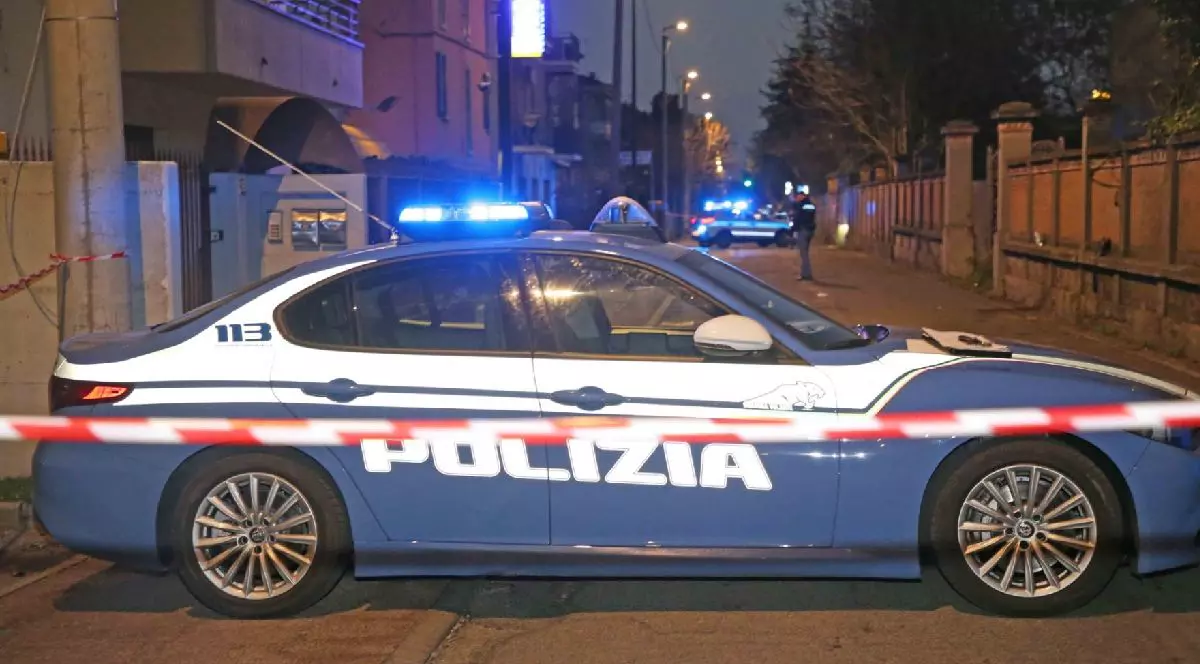 Fotografie cu o mașină de poliție italiană, cu lumini albastre intermitente, poziționată pe o stradă noaptea, izolată cu bandă roșie și albă.