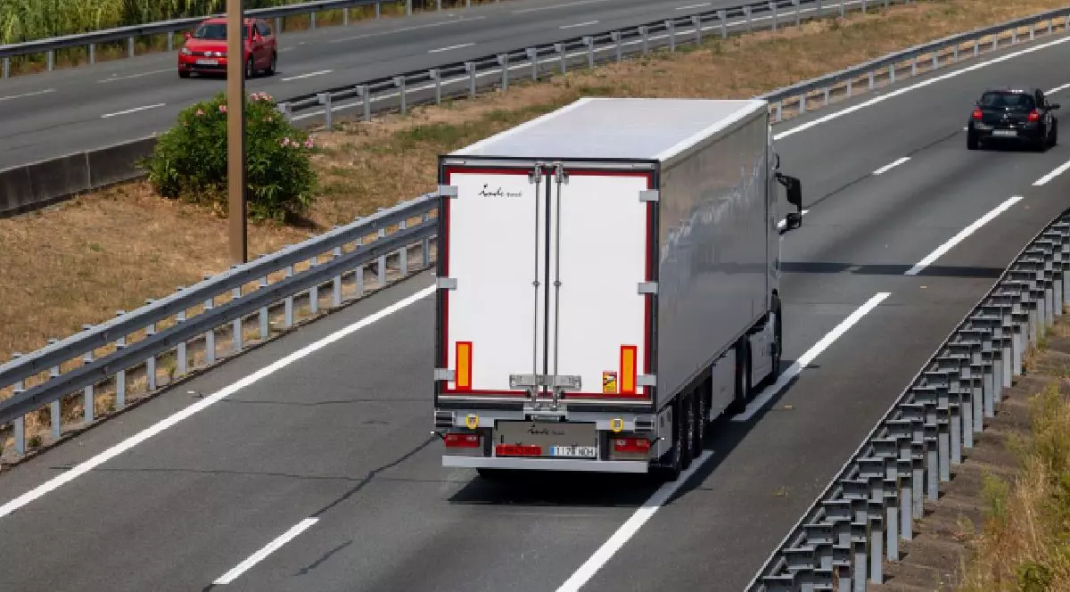 Fotografie cu un camion alb pe o autostradă cu mai multe benzi, cu parapete de protecție, o mașină neagră în fața și una roșie pe sensul opus.