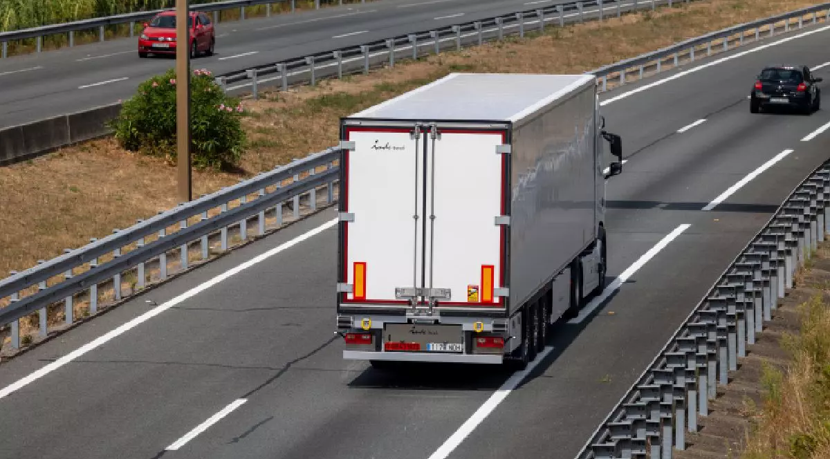 Fotografie cu un camion alb pe o autostradă cu mai multe benzi, cu parapete de protecție, o mașină neagră în fața și una roșie pe sensul opus.