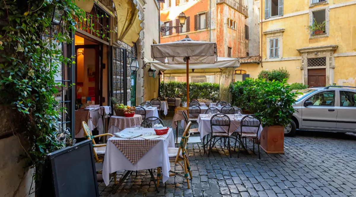 O stradă cu restaurante din Roma, mesele au scaune negre și fețe de masă albe, clădirile istorice au geamuri cu obloane