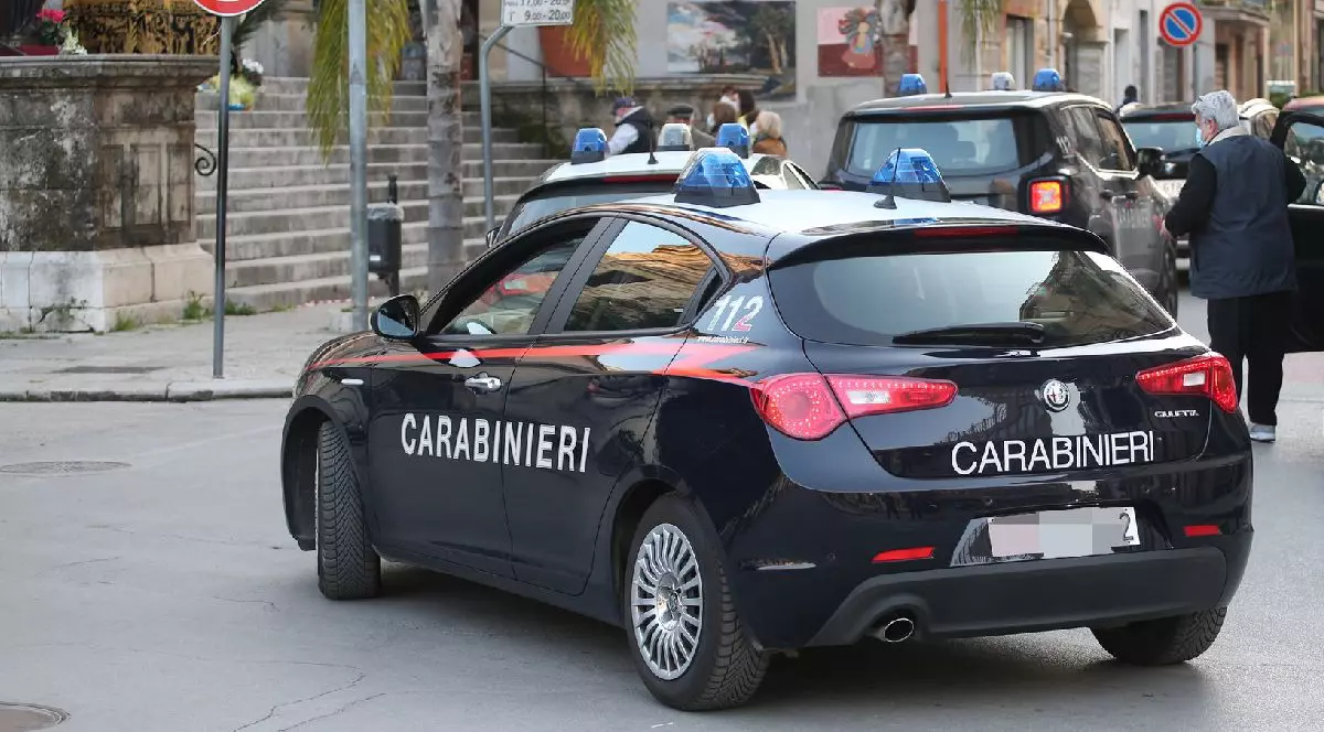 În imagine este o mașină neagră de poliție pe scrie CARABINIERI, staționată pe o stradă aglomerată, în fața unei clădiri, cu un semafor și indicatoare rutiere în fundal.