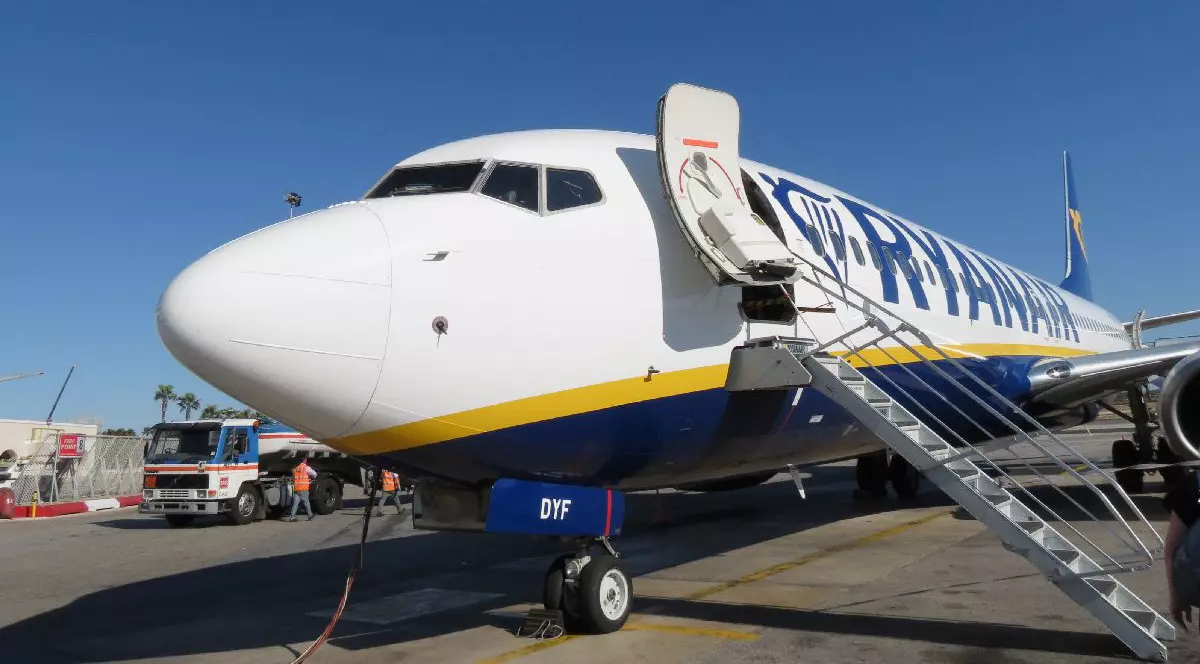 Fotografie cu un avion alb și albastru al companiei Ryanair, cu scara lăsată, într-un aeroport, sub un cer albastru senin.