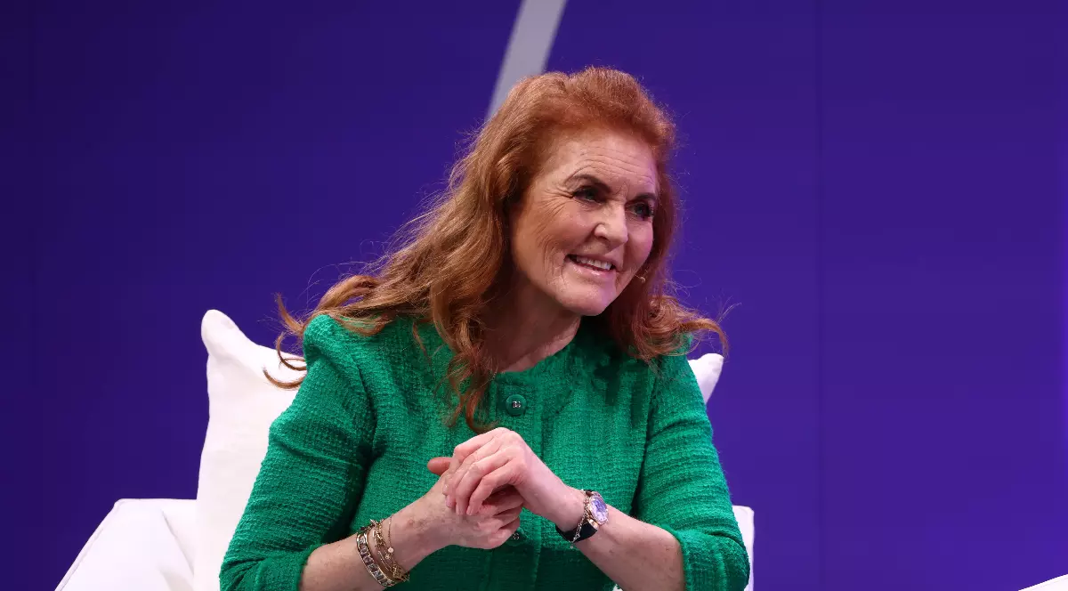 O femeie cu păr roșcat, îmbrăcată într-un costum verde elegant, stă așezată pe un fotoliu alb și zâmbește în timpul unei discuții. Fundalul este mov, iar ea poartă bijuterii discrete și ține câteva foi în mână.