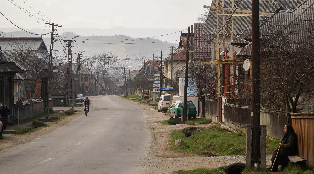 O stradă cu case dintr-un sat din România. Fiecare casă are gardul din lemn și bănci la poartă. Pe una dintre băci stă o femeie în vârstă, îmbrăcată în negru