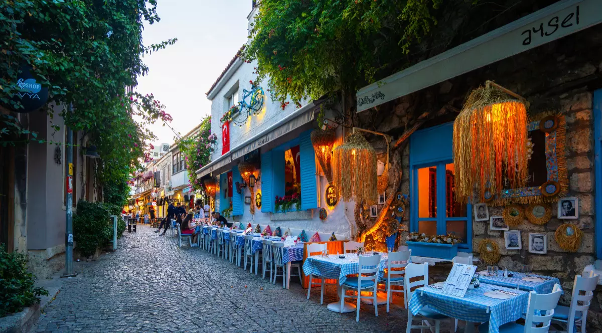 O stradă plină de restaurante din Alacati, Turcia. Maesele si scaunele albastre stau pe strada pietruita si sunt acoperite de fete de masă albe