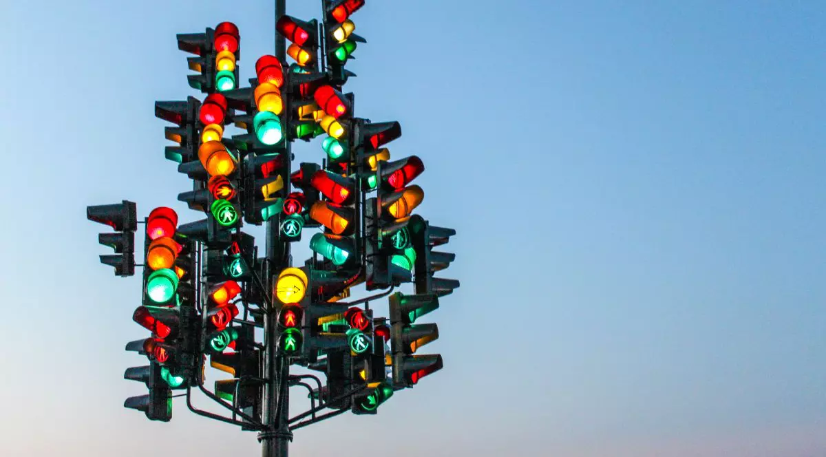Imagine creativă cu o mulțime de semafoare cu culorile roșu, galben și verde aprinse, pe un stâlp, iar pe fundal cerul albastru
