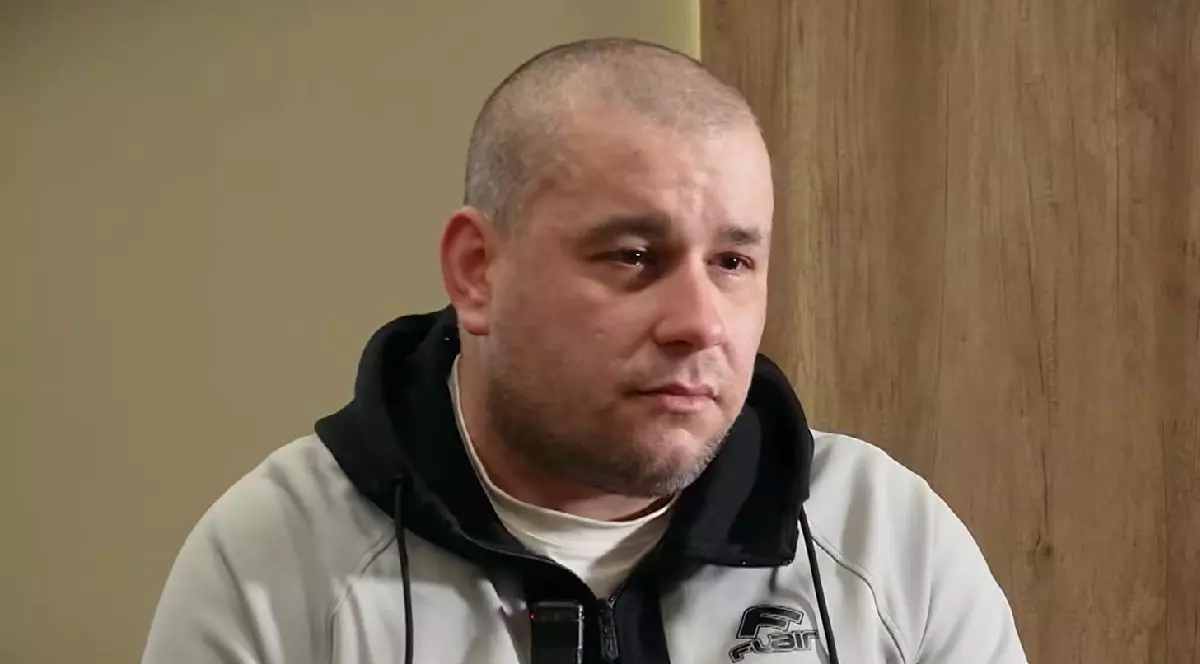 Un bărbat chel, cu o expresie serioasă, îmbrăcat într-o jachetă gri cu negru, cu lacrimi în ochi, stă în fața unui perete bej și a unei uși din lemn.