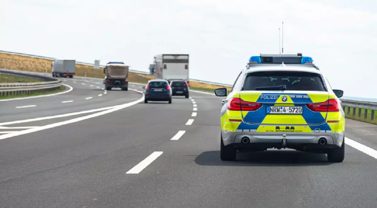 Imaginea arată o autostradă din Germania, având în prim-plan o mașină de poliție în mișcare. În fața autospecialei se află câteva autoturisme și trei camioane.