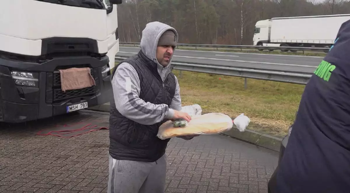 Un bărbat îmbrăcat într-un hanorac gri cu glugă și vesta neagră ține în mâini o pâine, într-o parcare, lângă un camion alb.