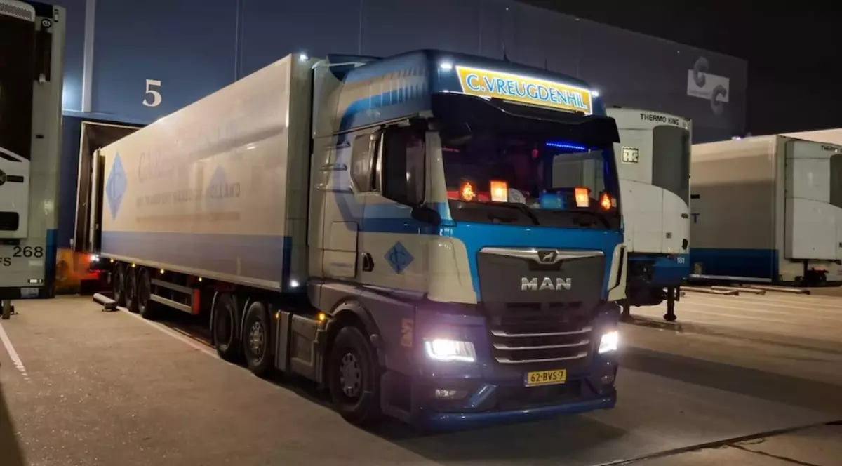 Fotografie surprinsă noaptea cu un camion MAN albastru parcat. Farurile și stopurile camionului sunt aprinse. Fundalul include un depozit slab iluminat.