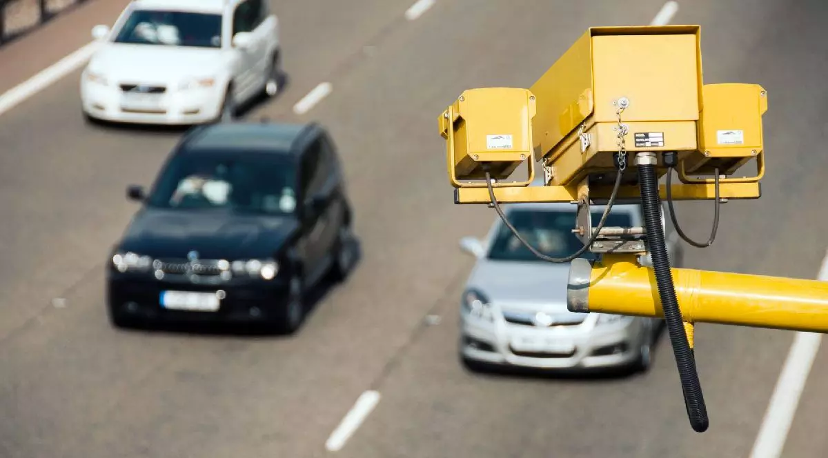 Vedere de sus a unei autostrăzi cu mai multe benzi. În prim-plan, un dispozitiv galben de monitorizare a traficului este montat pe un stâlp metalic. Trei mașini, un sedan negru, un hatchback argintiu și un sedan alb circulă în același sens, pe banzi diferite.