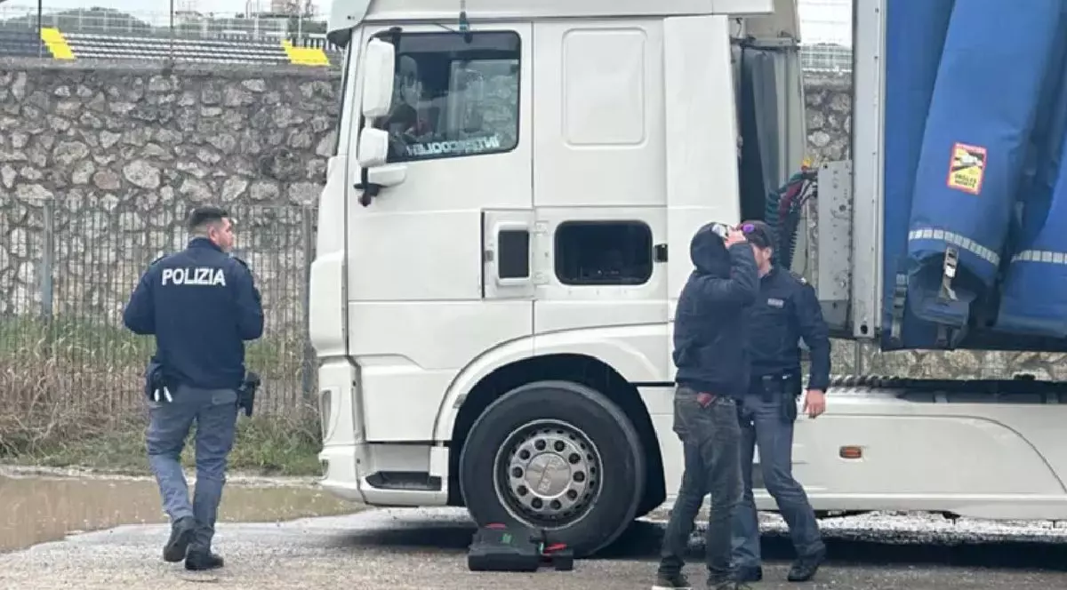 Fotografia arată trei bărbați, dintre care doi sunt polițiști, lângă un camion alb. În fundal se vede un zid de piatră și un gard metalic.
