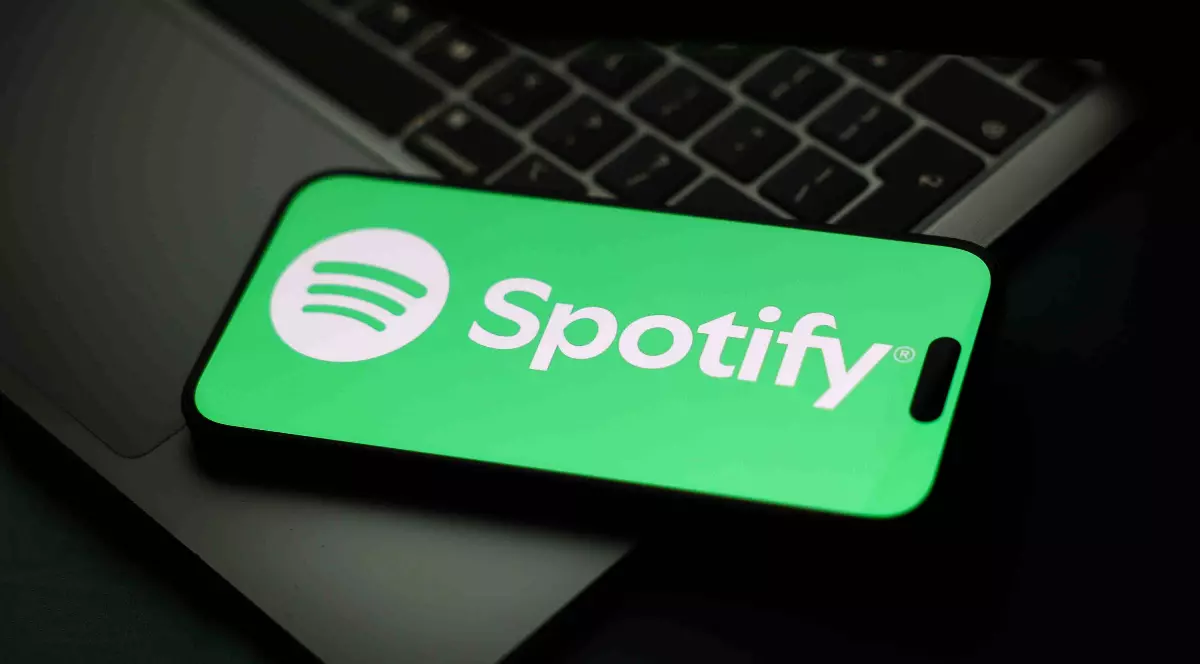 În imagine se vede un telefon mobil care afișează logo-ul Spotify pe ecran, așezat peste o tastatură de laptop. Fundalul întunecat scoate în evidență culoarea verde a aplicației, sugerând un context legat de tehnologie și muzică digitală.