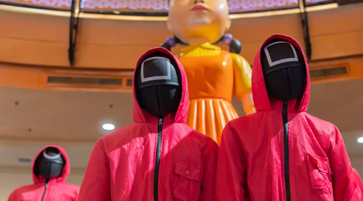 În imagine apar mai mulți gardieni îmbrăcați în costume roșii, cu măști negre, inspirați din serialul Squid Game.În spatele lor se vede celebra păpușă uriașă din joc, care accentuează atmosfera de control, supraveghere și tensiune.