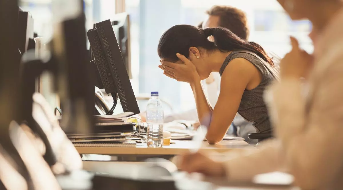 O tânără cu părul prins în coadă cu capul în mâini la birou, în fața unui monitor. Alte persoane estompate se află în apropiere