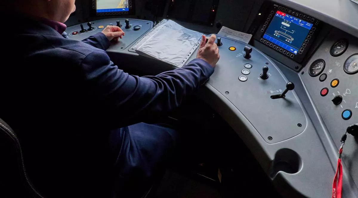 Imagine din interiorul unui tren de metrou , mecanicul este asezat si tine mana pe maneta, are in fata niste foi in folie de plastic