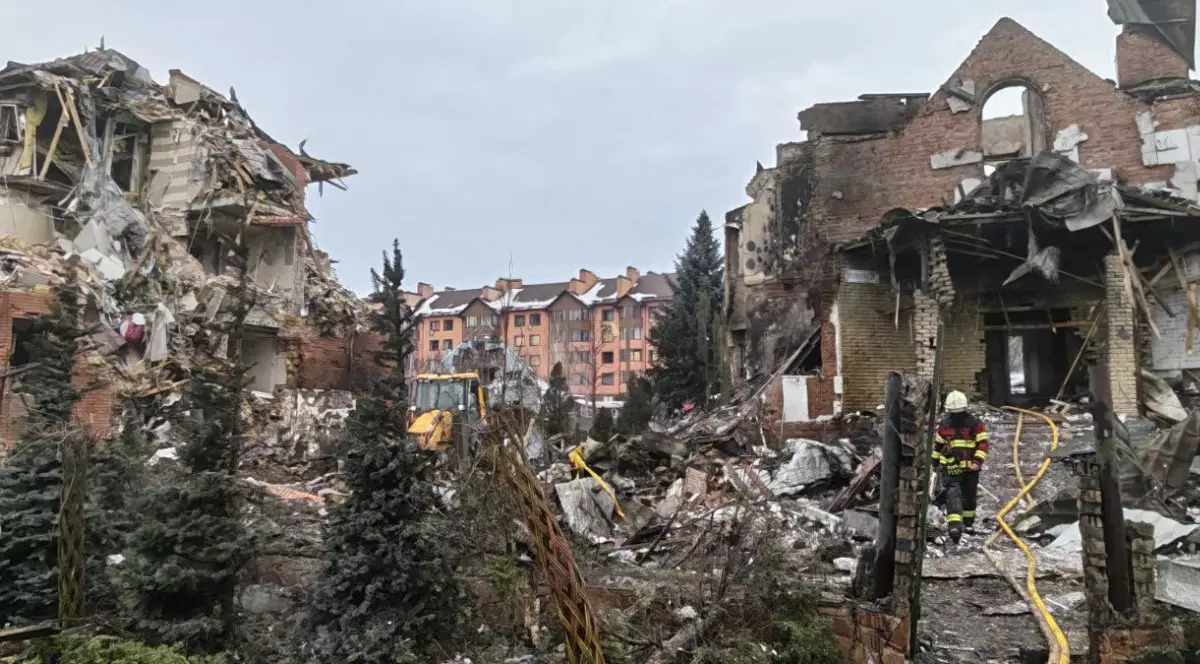 o scenă de distrugeri masive în exterior, unde două clădiri din cărămidă, situate în stânga și în dreapta cadrului, sunt parțial prăbușite, lăsând la vedere structurile interioare de lemn și beton. Spațiul dintre ele este acoperit de un morman mare de moloz compus din resturi de zidărie, grinzi rupte și bucăți de izolație, iar în centrul acestui perimetru se află un buldoexcavator galben parțial vizibil, Borșceahivka, 23 februarie 2026