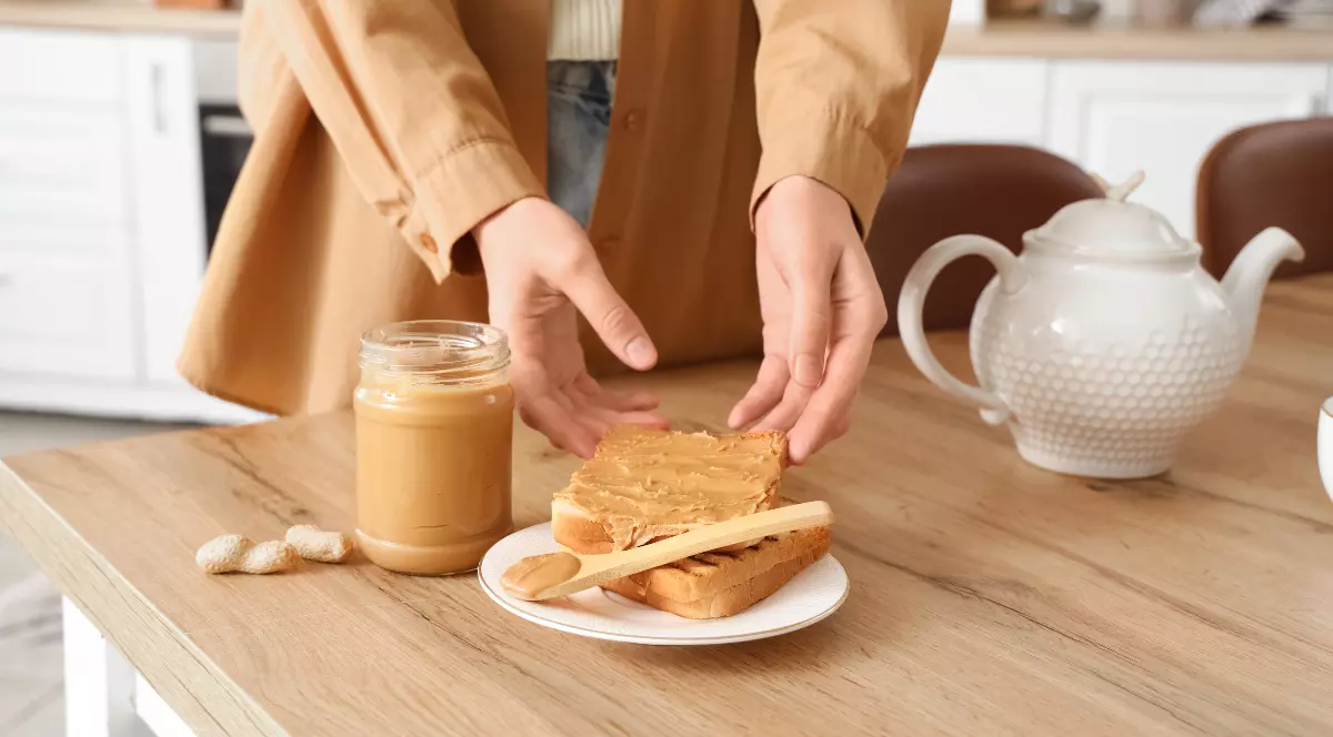 O persoană întinde unt de arahide pe o felie de pâine prăjită, într-o bucătărie luminoasă. Pe masă se află un borcan deschis cu unt de arahide, câteva arahide și un ceainic alb.
