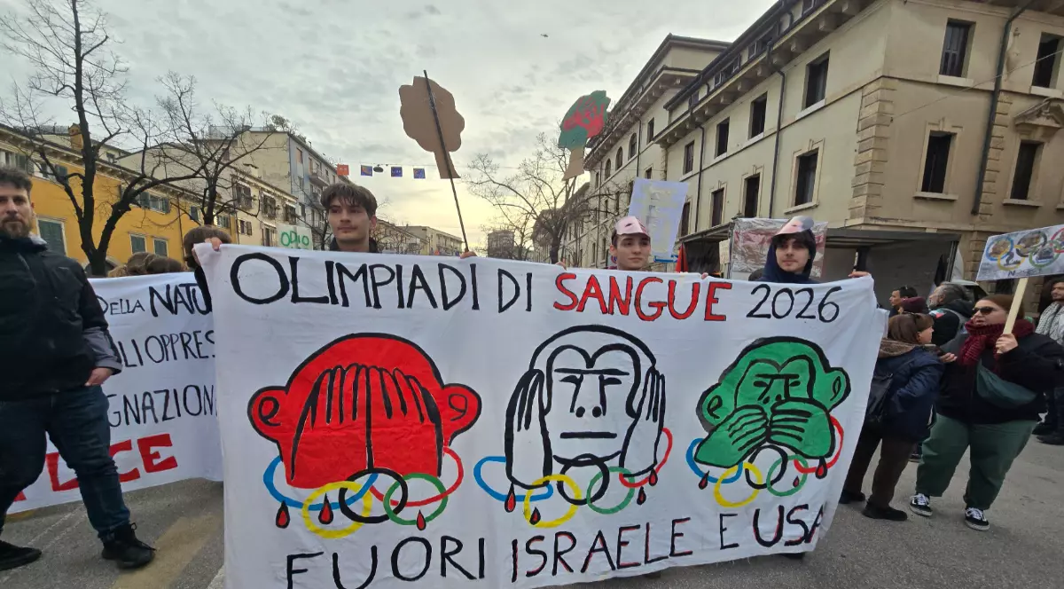 În imagine, mai mulți protestatari mărșăluiesc pe o stradă din Verona, ținând un banner mare pe care scrie „Olimpiadi di sangue 2026” și „Fuori Israele e USA”. Pe pancartă sunt desenate trei maimuțe stilizate, asociate cu simbolurile olimpice, într-un mesaj critic la adresa Jocurilor Olimpice.