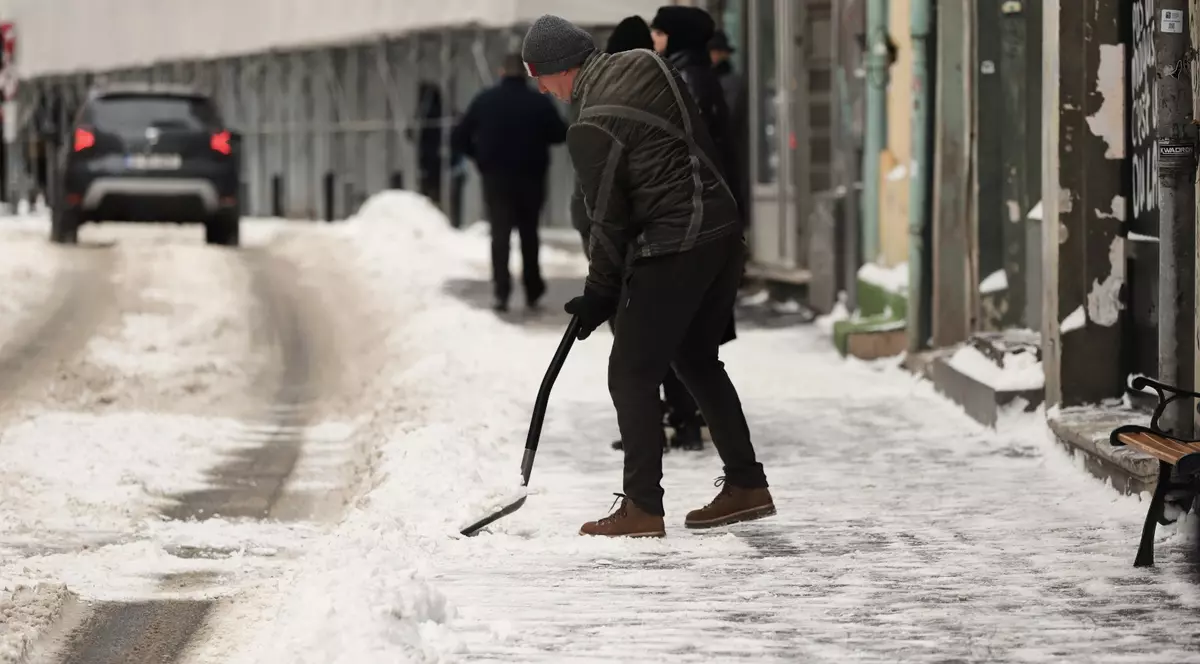 un bărbat îmbrăcat cu o jachetă matlasată gri-închis, pantaloni negri, ghete maro și o căciulă gri este aplecat în timp ce curăță zăpada de pe trotuar cu o lopată neagră. Trotoarul și marginea drumului sunt acoperite de un strat de zăpadă albă, parțial topită și murdară