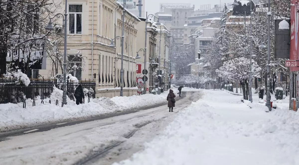 O stradă înzăpezită din București, cu clădiri acoperite de zăpadă, pietoni îmbrăcați în haine de iarnă.