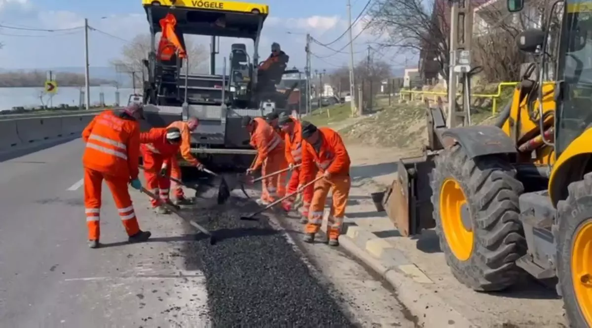 Mai mulți muncitori îmbrățai în costume portocalii lucrează la asfaltarea unei porțiuni de drum în Craiova, martie 2026