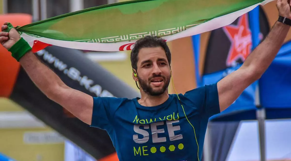 Amir Kiarash, singurul jurnalist român de origine iraniană, surprins în timpul unui eveniment sportiv. Ține deasupra capului un steag al Iranului, întins între ambele mâini ridicate. Zâmbește larg