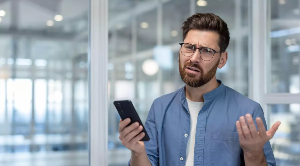 Un bărbat cu tricou alb, cămașă albastră, care ține un telefon în mână și este confuz