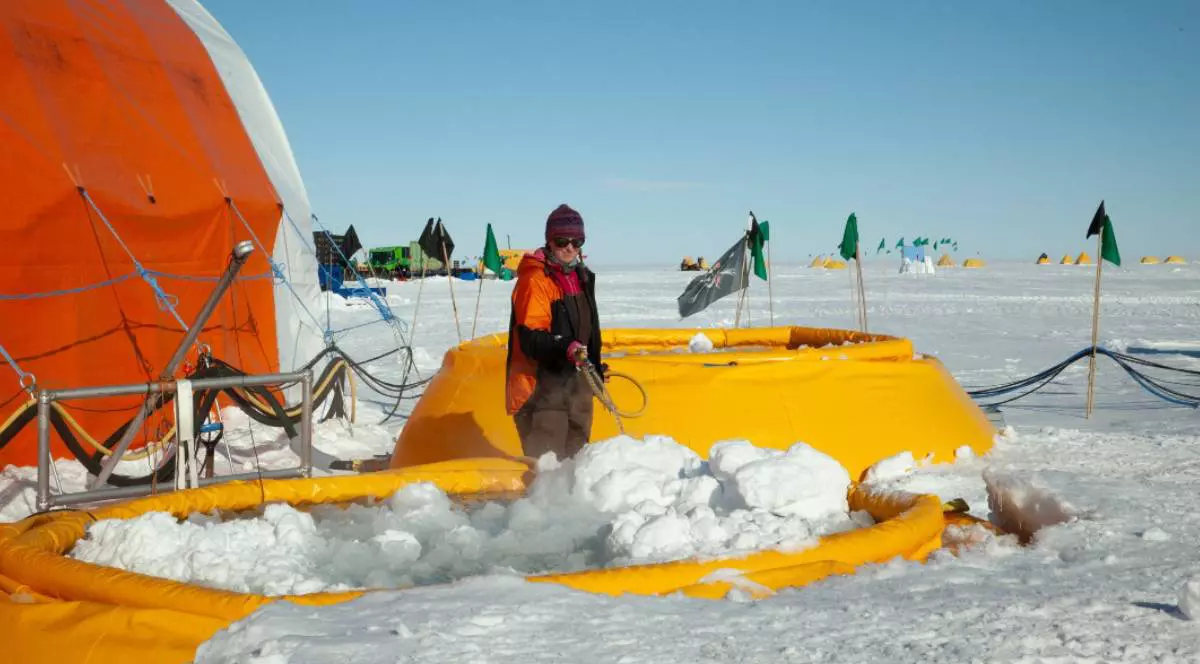 Un cercetător îmbrăcat în echipament gros de iarnă, portocaliu și negru, stă pe gheața din Antarctica și folosește un furtun care pulverizează apă fierbinte peste zăpadă. În jur se văd structuri gonflabile galbene, echipamente științifice și cabluri întinse. În fundal apar mai multe corturi colorate, dispuse pe câmpul alb de gheață, sub un cer senin.