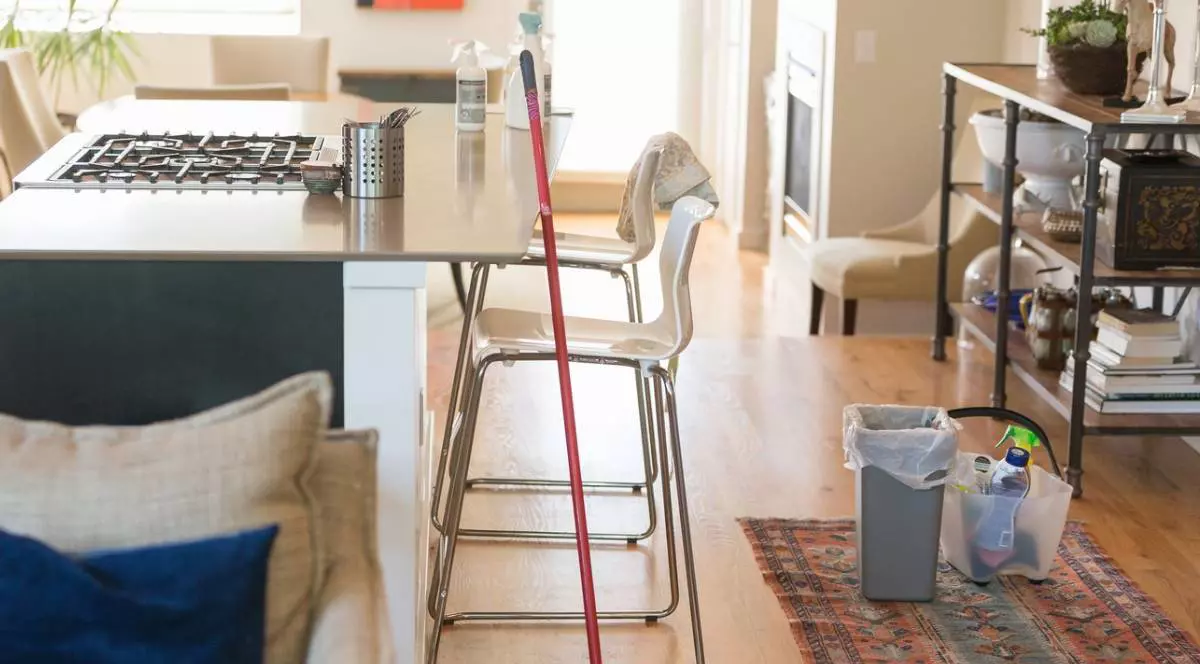 Imagine din interiorul unei case afalată în curățenie generala, camera este luminoasa, se vede un mop, pe jos o galeata cu produse de curatenie, alte cateva pe masa