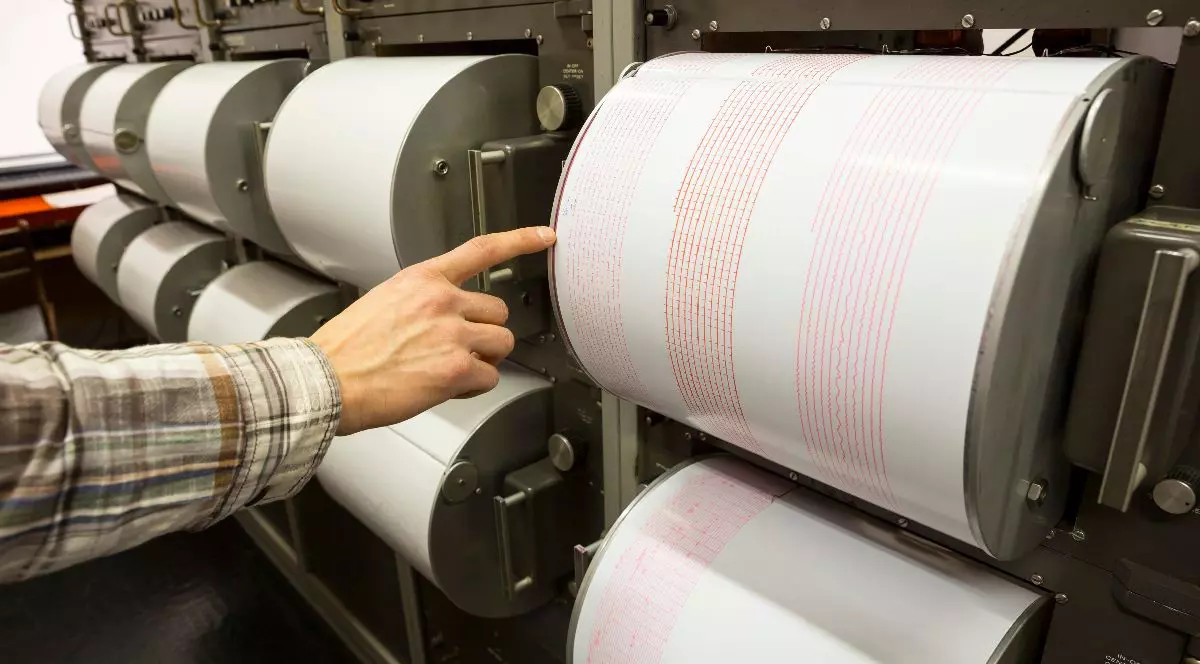 O fotografie în prim-plan arată o persoană îmbrăcată într-o cămașă în carouri, care arată spre un seismograf.