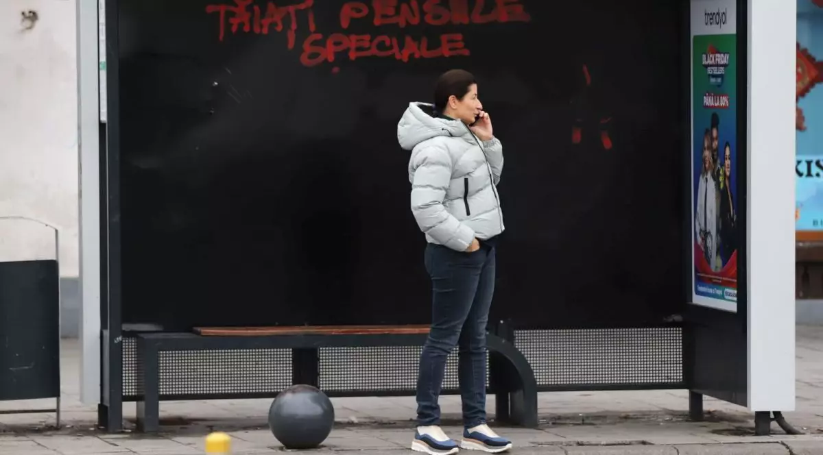 O femeie este în stația de autobuz și vorbește la telefon, iar pe panoul din spatele ei scrie, roșu pe negru Tăiați pensiile speciale, în București, ianuarie 2026