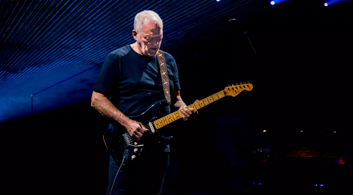 Imaginea îl surprinde pe un chitarist cântând pe scenă, sub lumini albastre puternice și efecte vizuale de concert. Artistul, îmbrăcat într-un tricou negru, este concentrat asupra chitarei electrice, în timp ce atmosfera spectacolului este accentuată de luminile și grafica din fundal.