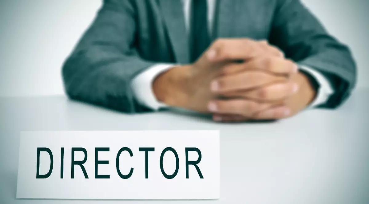 un bărbat în costum, așezat la un birou, cu o plăcuță cu numele în fața lui pe care scrie „director”