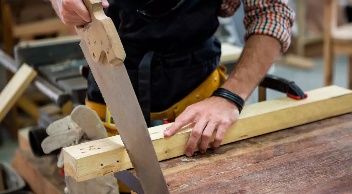 Un bărbat lucrează într-un atelier de tâmplărie și taie o bucată de lemn cu un fierăstrău manual mare. Ține lemnul cu o mână, iar cu cealaltă mișcă fierăstrăul înainte și înapoi. Poartă o cămașă în carouri și un șorț de protecție negru. Pe masă se văd alte unelte și bucăți de lemn, iar în fundal apare atelierul, cu mobilier și materiale specifice lucrului cu lemnul.