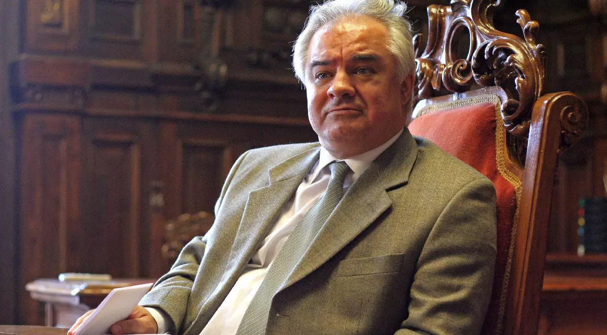 O imagine cu Eugen Rădulescu, consilier BNR, îmbrăcat într-un costum gri și cravată, așezat la o masă masivă de lemn într-un decor clasic, cu mobilier sculptat. Ține în mână câteva foi de hârtie.