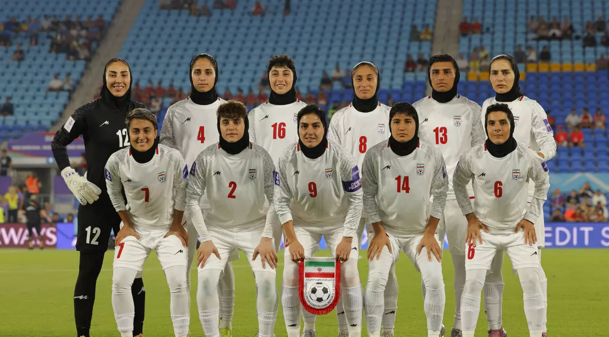 Echipa națională feminină de fotbal a Iranului pozează pe teren înaintea unui meci, aliniată pentru fotografia oficială. Jucătoarele poartă echipament alb și hijab, iar în fundal se pot observa tribunele stadionului.